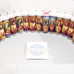 May include: A collection of 20 colorful prayer candles featuring various people's faces. The candles are arranged in a semi-circle on a white surface. The candles are all the same size and shape, and they have a white wax base with a colorful image on the front. The images are all of people's faces, and they are all in a similar style. The candles are all lit, and they are casting a warm glow on the surface.