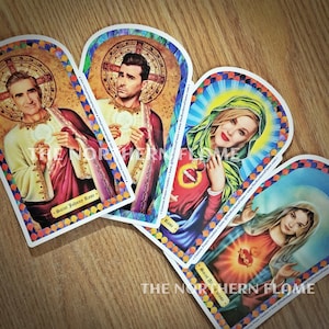 May include: Four colorful prayer candle holders with arched tops. Each holder features a different saint, including Saint Johnny Rose, Rose, Saint Alis Mose, and a fourth saint with a heart in their hand. The holders are decorated with colorful stained glass designs and have the text "Custom Prayer Candles" and "The Northern Flame" printed on them.