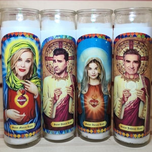 May include: Four white pillar candles with colorful images of people and the text "Saint Moira Rose", "Saint David Rose", "Saint Alexis Rose", and "Saint Johnny Rose".