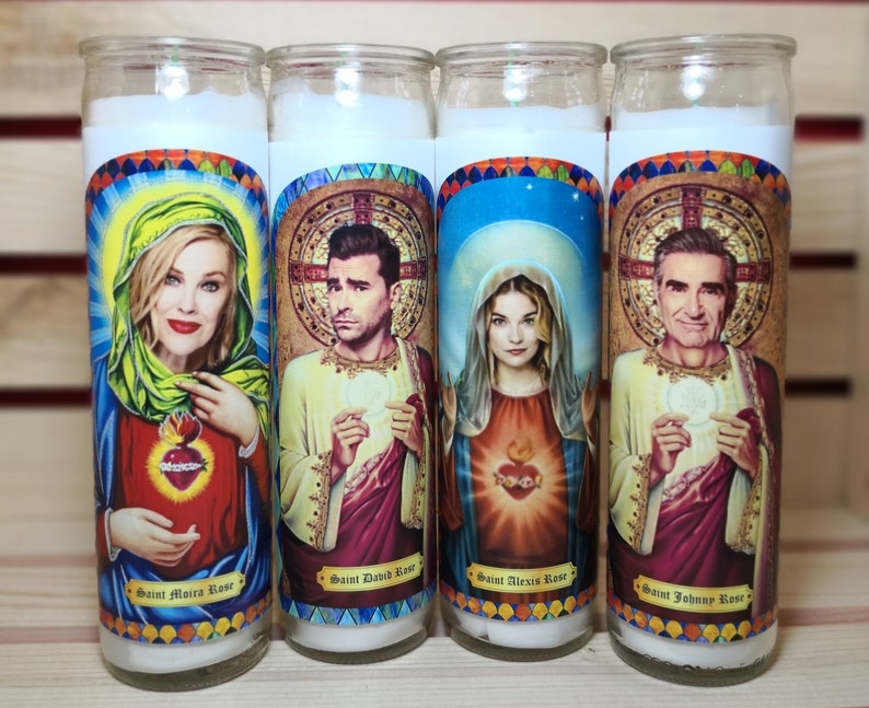 May include: Four white pillar candles with colorful images of people and the text "Saint Moira Rose", "Saint David Rose", "Saint Alexis Rose", and "Saint Johnny Rose".