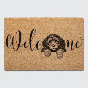 May include: A brown coir doormat with the word "welcome" in black cursive script. A black and white illustration of a dog peeking over the top of the mat.