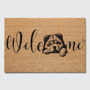 May include: A brown coir doormat with the word "welcome" in black cursive script. A black and white illustration of a Chow Chow puppy peeking over the top of the mat.