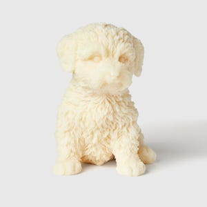 May include: A white, fluffy, sitting puppy candle. The candle is made of wax and has a realistic appearance.