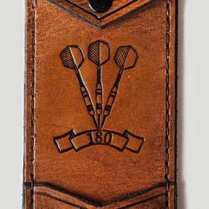 May include: A brown leather dart case with a flap closure and a snap button. The case has a pocket on the front with the initials "E.G." and a dart design with the number "180" underneath.