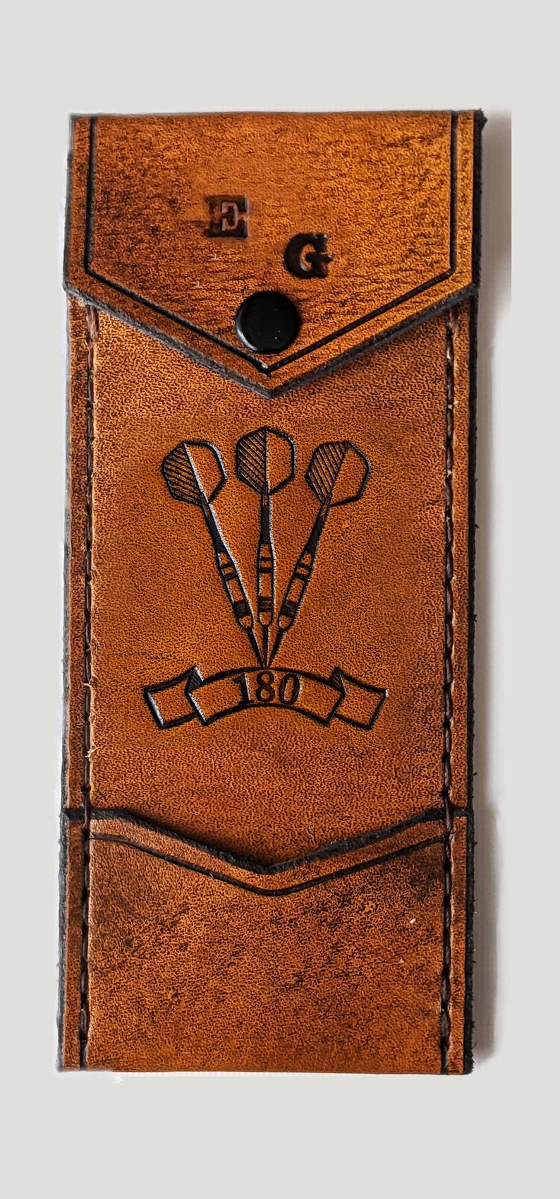 May include: A brown leather dart case with a flap closure and a snap button. The case has a pocket on the front with the initials "E.G." and a dart design with the number "180" underneath.