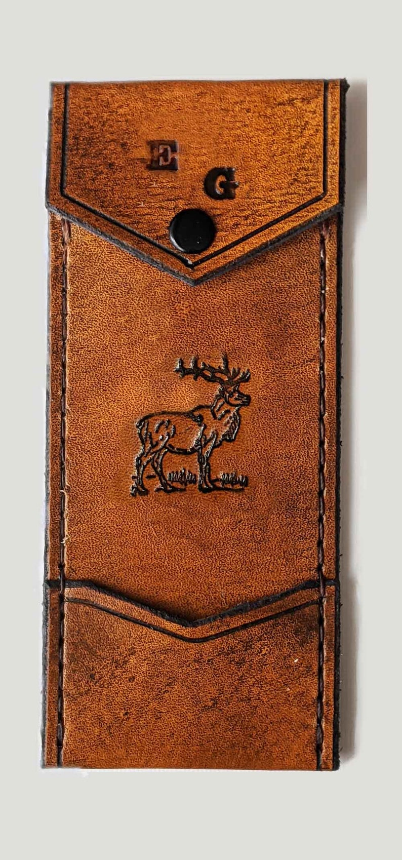 May include: A brown leather wallet with a flap closure and a snap button. The wallet has a pocket on the front and a deer silhouette embossed on the front. The initials "E.G." are embossed on the top flap.