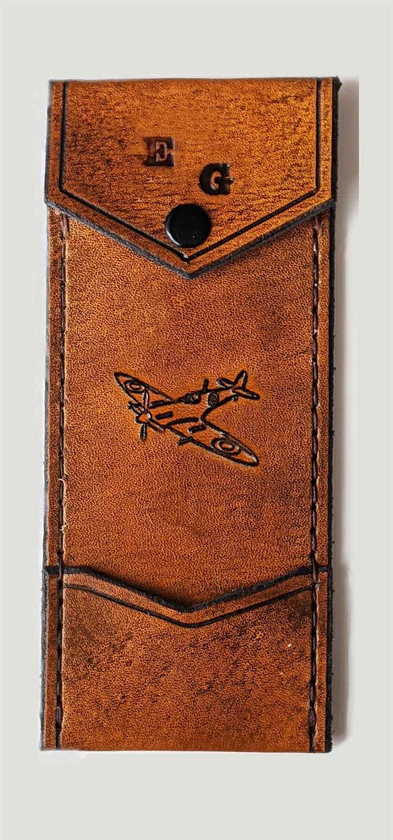 May include: A brown leather wallet with a flap closure and a snap button. The wallet has a pocket on the front with the initials "E.G." embossed on it. A small airplane is embossed on the front of the wallet.