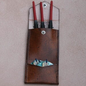 May include: A brown leather dart case with three red darts and a set of blue and white dart flights inside. The case has a flap with the text "Genuine Leather Handcrafted" printed on it.