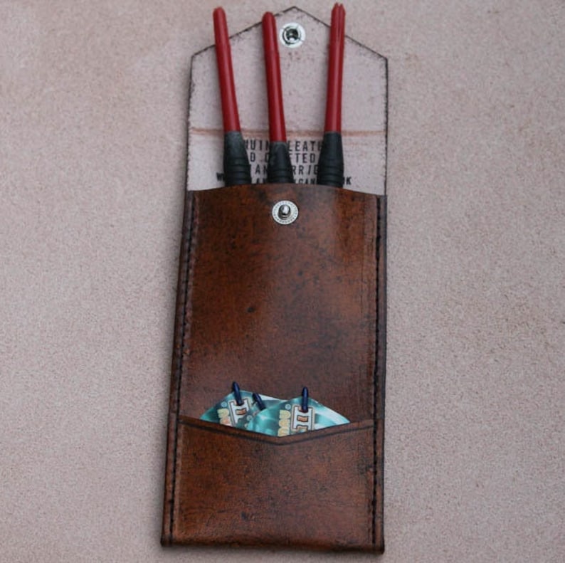 May include: A brown leather dart case with three red darts and a set of blue and white dart flights inside. The case has a flap with the text "Genuine Leather Handcrafted" printed on it.