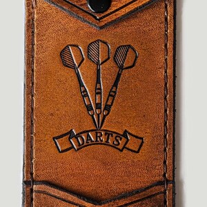 May include: A brown leather dart case with a black snap closure. The case has a pocket on the front with the initials "E.G." and the word "DARTS" embossed on it.