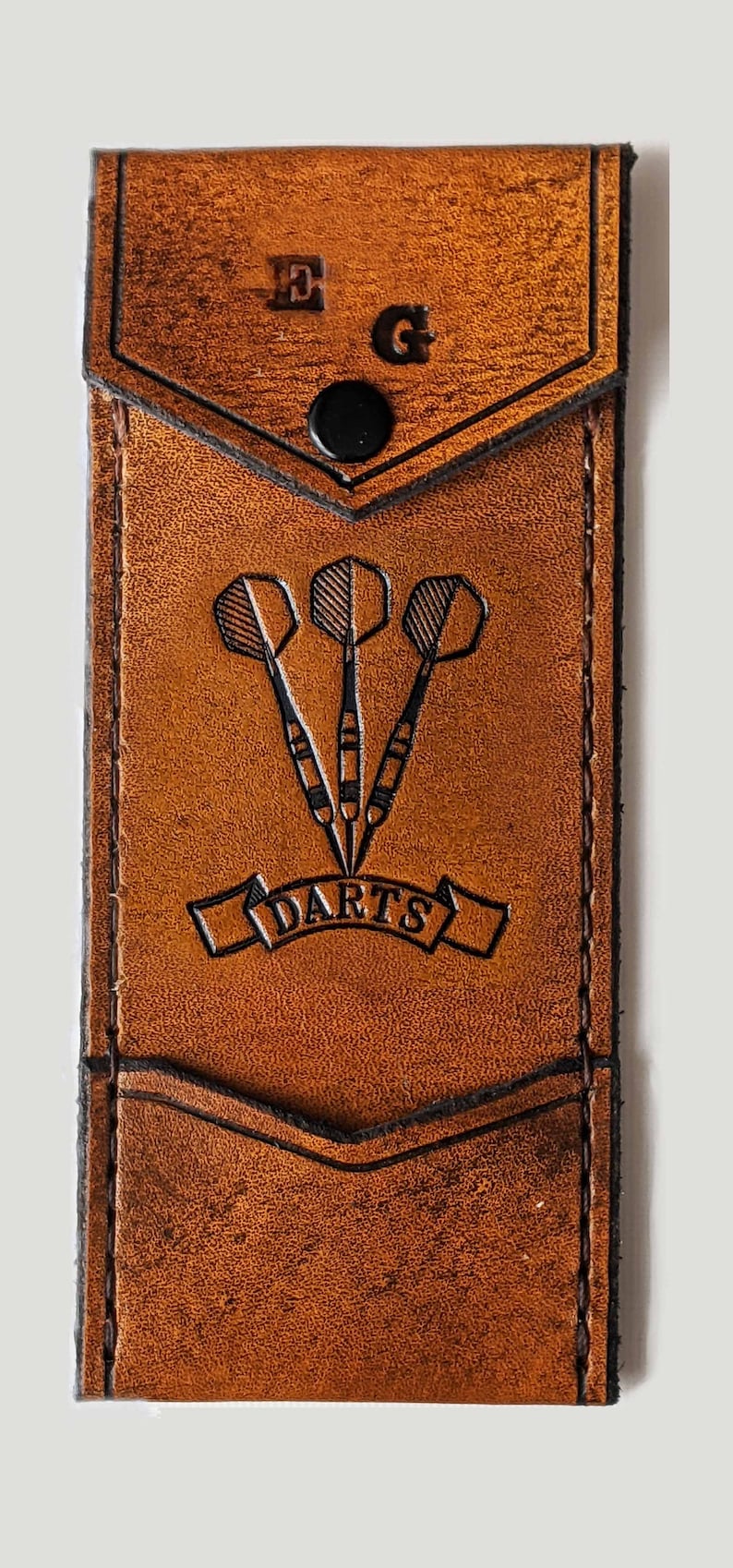 May include: A brown leather dart case with a black snap closure. The case has a pocket on the front with the initials "E.G." and the word "DARTS" embossed on it.
