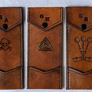 May include: Three brown leather cases with different designs. The first case has a skull and crossbones design and the initials "SA". The second case has a Celtic knot design and the initials "GH". The third case has a dartboard design with three darts and the initials "ES".