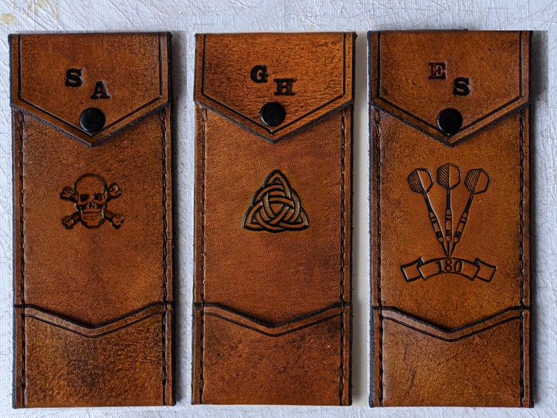 May include: Three brown leather cases with different designs. The first case has a skull and crossbones design and the initials "SA". The second case has a Celtic knot design and the initials "GH". The third case has a dartboard design with three darts and the initials "ES".