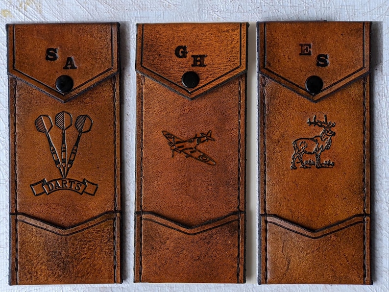May include: Three brown leather pouches with different designs. The first pouch has a dart design and the initials "SA". The second pouch has a plane design and the initials "GH". The third pouch has a deer design and the initials "ES".