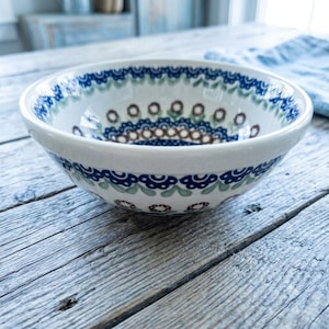 May include: A round ceramic bowl featuring a cream-colored base with a decorative pattern of dark blue scalloped edges, light green leaf motifs, and small brown circular accents arranged in concentric rings around the interior and exterior.