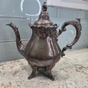 May include: An antique silver teapot with ornate detailing. The teapot features a curved spout, a decorative handle, and rests on three small feet. The lid is topped with a decorative finial. The teapot has a vintage aesthetic.