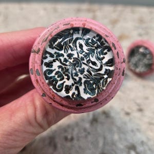 Pode incluir: Um puxador redondo com uma borda externa rosa salpicada e uma incrustação central de vidro. A incrustação exibe um padrão floral de damasco detalhado em preto e branco. Um segundo puxador correspondente é visível ao fundo.