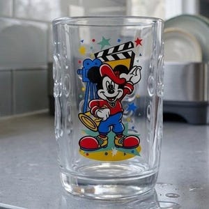 May include: A clear glass tumbler featuring a colourful graphic of Mickey Mouse dressed in a red shirt, blue shorts, and red shoes. Mickey is positioned in front of a blue film camera and a black and white film clapperboard, surrounded by small stars.