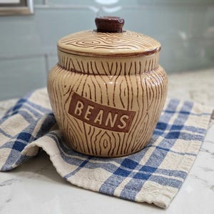 May include: A ceramic canister with a tan base and brown wood grain pattern. The word BEANS is embossed on the front in a rectangular panel. It features a matching lid with a dark brown knob handle, designed for kitchen storage and organization.