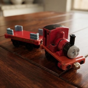 May include: A red toy train engine with a grey face and black chimney attached to a red flatbed cargo car. The flatbed car features light blue side panels and the text Sodor Shipping printed in yellow on the side of the red frame.