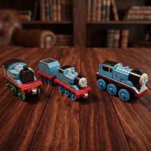 May include: Three toy trains on a wooden surface. The left train is blue with a black front and red wheels. The middle train is blue with the number 2 on a tender and the number 1 on the engine. The right train is blue with the number 1 on the side.