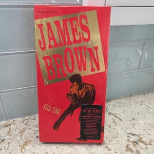 Könnte beinhalten: Eine rote Compact-Disc-Edition der "Star Time"-Sammlung von James Brown. Die Box hat goldfarbene Schriftzüge und ein goldenes Quadrat mit dem Namen "JAMES BROWN". Auf der Vorderseite befindet sich eine Schwarz-Weiß-Illustration von James Brown.