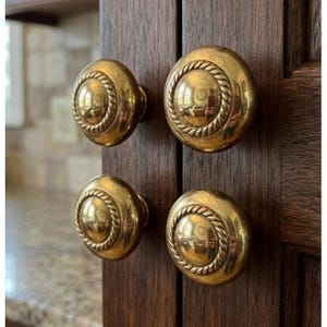 Puede incluir: Cuatro pomos redondos de latón para armarios con acabado pulido y un detalle decorativo de borde estilo cuerda. Cada pomo está montado en una puerta de armario de madera de color marrón oscuro, con una superficie lisa y reflectante.