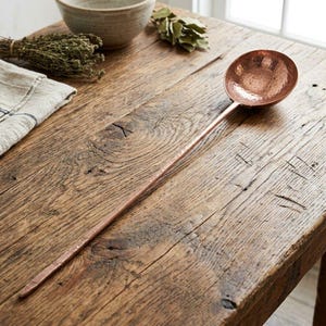 May include: A long-handled copper ladle featuring a round, hammered bowl and a slender, textured stem. The metal surface displays a warm, metallic reddish-brown finish with visible indentations from the hammering process along the handle.