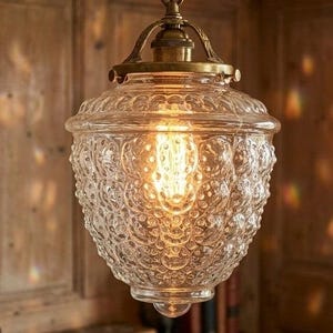 May include: A pendant light fixture featuring a clear, textured glass shade with a repeating embossed pattern. The shade is secured by a metallic brass-toned hardware mount at the top, which holds a glowing, warm-toned Edison-style light bulb inside.