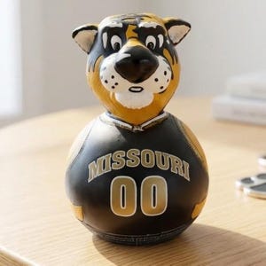 Puede incluir: Una figura de calabaza pintada a mano con forma de mascota de tigre. El tigre tiene una cara amarilla y negra, y el cuerpo está pintado como una camiseta negra con la palabra MISSOURI y el número 00 en letras doradas con bordes blancos.