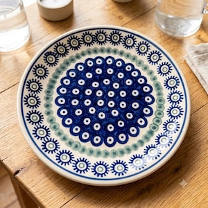 May include: A round ceramic plate featuring a cream-coloured base with a dense central pattern of dark blue circles, surrounded by a ring of soft green dots and an outer border of dark blue sunburst-like motifs with small white centres.