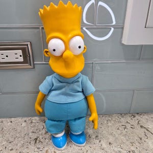 May include: A Bart Simpson doll with a yellow head and spiky hair, large white eyes, and a prominent nose. He wears a light blue shirt, blue pants, and blue shoes with white accents. The doll is standing on a countertop.