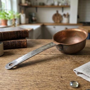 May include: A round copper ladle with a long, flat, weathered metal handle featuring a circular hole at the end. The bowl of the ladle has a warm, metallic copper finish, while the handle shows signs of age and oxidation with a dull gray patina.