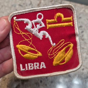 Puede incluir: Un parche bordado cuadrado con fondo rojo y borde blanco. Presenta una figura blanca estilizada que sostiene balanzas amarillas, el símbolo del zodiaco Libra en amarillo y el texto blanco LIBRA bordado en la parte inferior del parche.