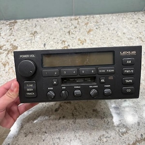 Puede incluir: Autorradio Lexus negro con botones para encendido, volumen, sintonización y selección de pistas. Incluye opciones de AM/FM, casete y reproductor de CD. Con controles de bajos, medios, agudos, balance y atenuador. El texto "THEFT DETERRENT DESIGN" es visible.