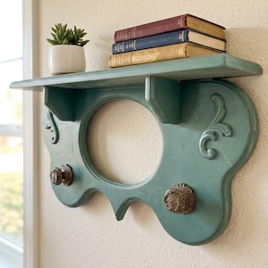 May include: A teal wall-mounted shelf with a central oval cutout, two ornate metal hooks, and decorative scrollwork. A small white pot containing a green succulent sits on the shelf, topped by a stack of three books with colored spines.
