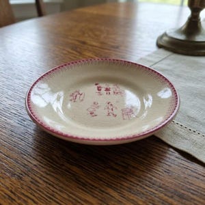 May include: A small, round, cream-colored ceramic saucer featuring a delicate red decorative border and a central pattern of small, red, line-drawn figures of children playing. The surface of the saucer displays a visible, fine crackle glaze finish.