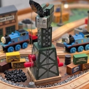 May include: A wooden toy crane with a smiling face, featuring a green and gray tower structure, red side cranks, and a hanging hook. The crane is surrounded by blue toy train engines, wooden crates labeled Sodor Steamworks, and a pile of black coal.