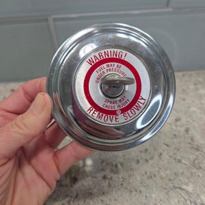 May include: A shiny, chrome-colored fuel cap with a red and white warning label. The label reads "WARNING! FUEL MAY BE UNDER PRESSURE. SPRAY MAY CAUSE INJURY. REMOVE SLOWLY." A small key-like lever is in the center.