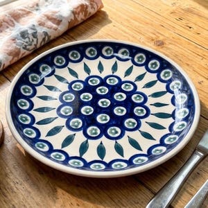 May include: A round ceramic plate featuring a cream-coloured base with a hand-painted pattern of dark blue circles containing small green flowers, surrounded by a ring of dark blue leaf shapes and an outer border of blue-rimmed green floral motifs.