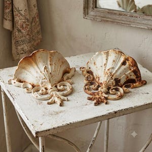 May include: Two wall-mounted half-shell planters made of off-white painted metal with visible brown rust accents. Each piece features a fluted shell design with ornate scrollwork at the base, creating a decorative semi-circular basin for wall display.