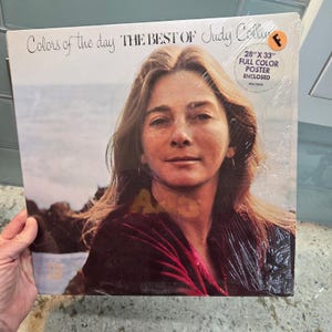 Puede incluir: Un álbum de vinilo titulado "Colors of the Day: The Best of Judy Collins". La portada del álbum presenta un retrato de Judy Collins. El álbum está sellado en plástico e incluye un póster a color de 71 cm x 84 cm.
