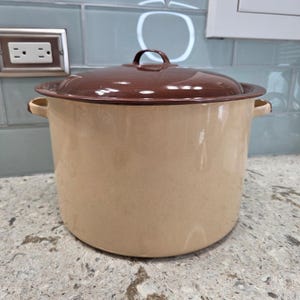 May include: A large, beige enamel pot with a brown lid and handle. The pot has two side handles and sits on a speckled countertop. The lid has a rounded handle. Kitchenware.
