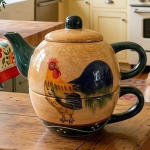 Peut inclure: Un service à thé en céramique pour une personne composé d'une théière empilée sur une tasse assortie. L'ensemble présente une finition émaillée craquelée beige et est orné d'une illustration de coq coloré, avec un bec, une anse et un bouton vert foncé.
