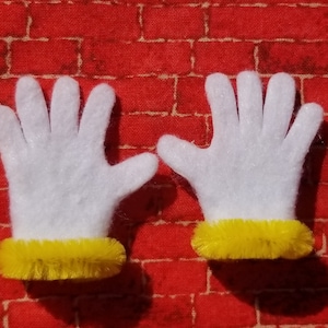 Könnte beinhalten: Ein Paar weiße Filzhandschuhe mit gelben, flauschigen Manschetten. Die Handschuhe liegen auf einem roten Ziegelhintergrund.