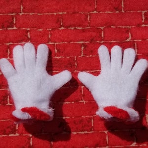 Könnte beinhalten: Ein Paar weiße Filzhandschuhe mit roten Filzmanschetten. Die Handschuhe liegen auf einem roten Ziegelhintergrund.