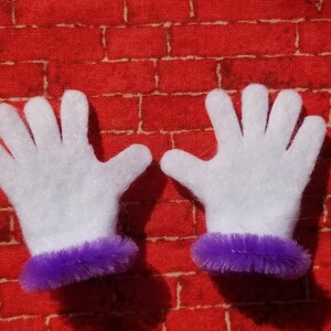 Könnte beinhalten: Ein Paar weiße Filzhandschuhe mit violetter, flauschiger Einfassung. Die Handschuhe liegen flach auf einem roten Ziegelhintergrund.