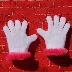 May include: A pair of white felt gloves with pink fur trim. The gloves are on a red brick background.