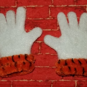 May include: A pair of white felt gloves with orange and black striped cuffs. The gloves are on a red brick background.