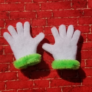 May include: A pair of white felt gloves with green fuzzy trim. The gloves are on a red brick background.